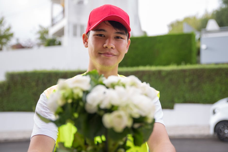 A male flower delivery courier wearing a red cap and a reflective vest is smiling as he holds out a bouquet of fresh white flowers, possibly roses and lilies, wrapped in clear plastic, towards the cam
