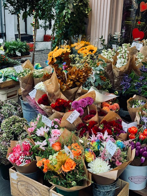 A vibrant outdoor flower display featuring an assortment of fresh flowers including bright yellow sunflowers, pink lilies, red and purple chrysanthemums, orange roses, white daisies, and purple irises