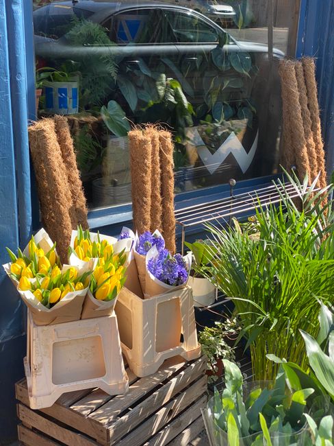 Outdoor display of potted flowers and floral arrangements at a florist shop near Hackney Wick station. The arrangement includes yellow tulips and purple flowers wrapped in brown paper, placed on woode