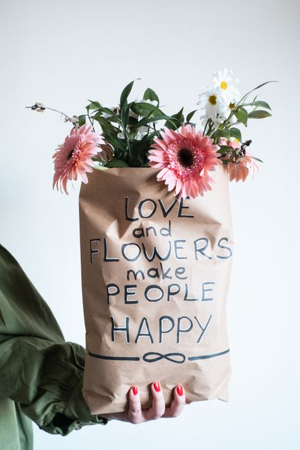 A person holds a brown paper bag floral gift with the handwritten message 'Love and Flowers make People Happy' in black and blue ink. The bouquet inside includes pink Gerbera daisies, white daisies wi