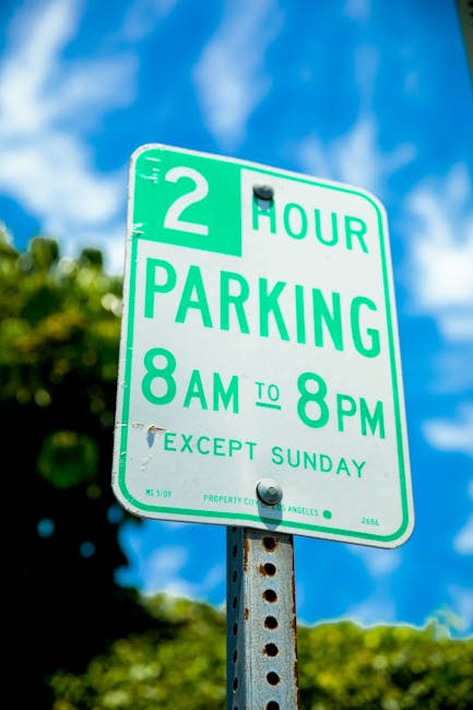 A close-up of a green and white parking regulation sign mounted on a weathered metal pole. The sign indicates a 2-hour parking limit from 8 am to 8 pm, Monday to Saturday, with exceptions for Sundays.