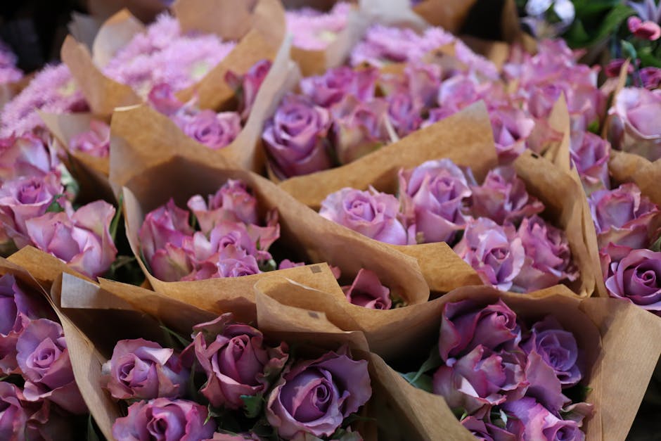 A close-up view of numerous small bouquets of fresh pink and lavender roses, wrapped in light brown kraft paper, arranged in a floral shop setting. The roses display delicate petals with slight variat