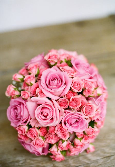 A round bouquet arrangement of fresh pink roses of varying sizes, with some still in bud form and others fully bloomed, featuring soft, velvety petals. The flowers are densely packed, creating a lush,