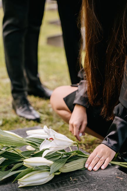 A person kneeling at a gravesite, placing a floral arrangement of white lilies with long green leaves onto a black headstone. The lilies have large, trumpet-shaped petals with some partially open, dis