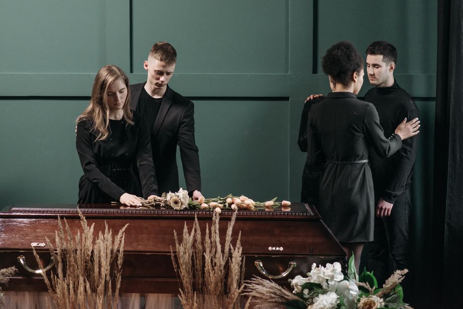Two women and two men dressed in black mourning attire stand around a closed wooden coffin decorated with a floral arrangement of white roses, pale pink tulips, and greenery, placed on a stand in a di